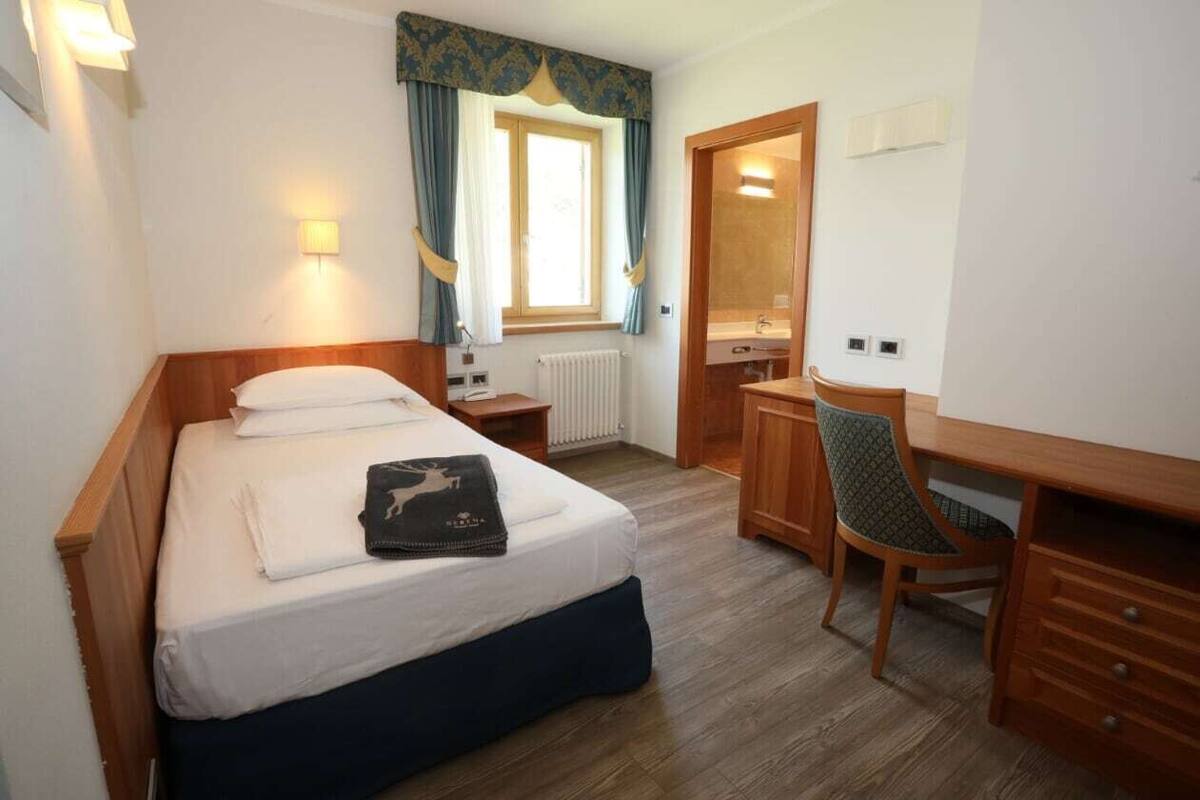 Une chambre d’hôtel comprenant un lit, une table de chevet, un bureau, une chaise et une fenêtre avec des rideaux.