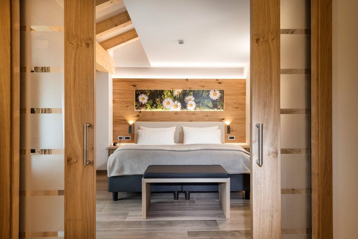 Une chambre à coucher moderne avec une tête de lit en bois, un grand lit avec une literie blanche, un banc au pied du lit et une œuvre d’art représentant des fleurs, accrochée au mur.