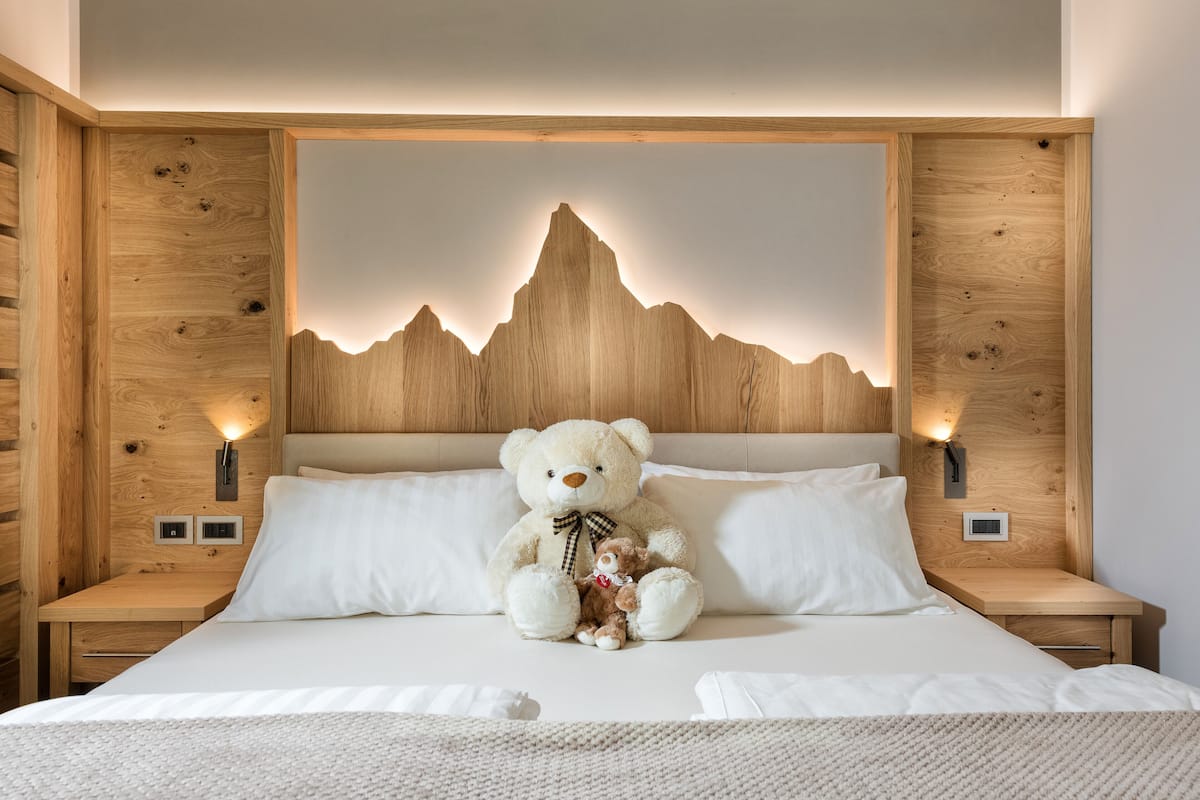 Un lit avec un grand ours en peluche et un plus petit animal en peluche, une tête de lit en bois ornée d’un motif de montagne et des lampes de chevet.