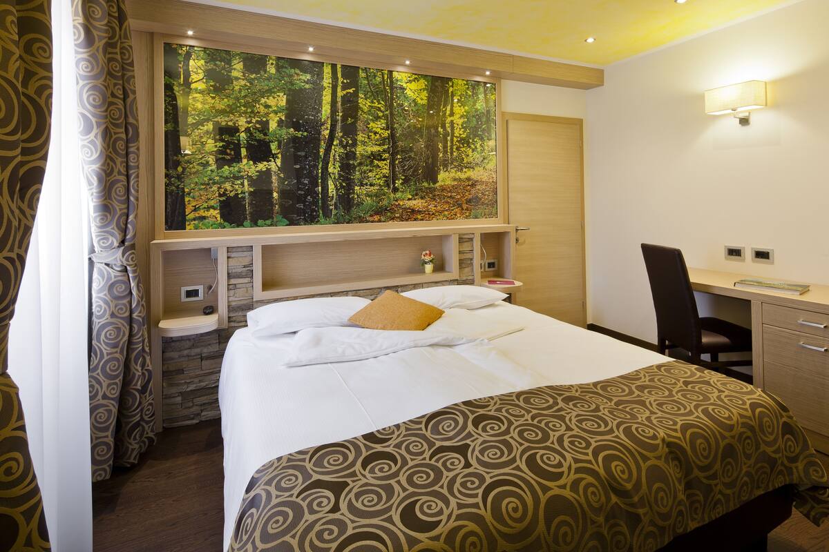 Une chambre d’hôtel comprenant un lit, un bureau, une chaise, une grande fenêtre donnant sur la forêt et un luminaire fixé au mur.