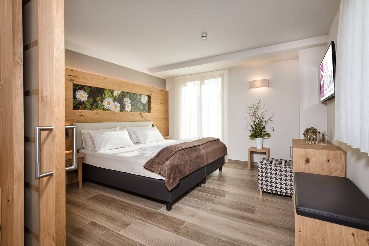 Une chambre à coucher moderne avec une tête de lit en bois, un grand lit, une table de chevet agrémentée d’une plante et un pouf à motif pied-de-poule.