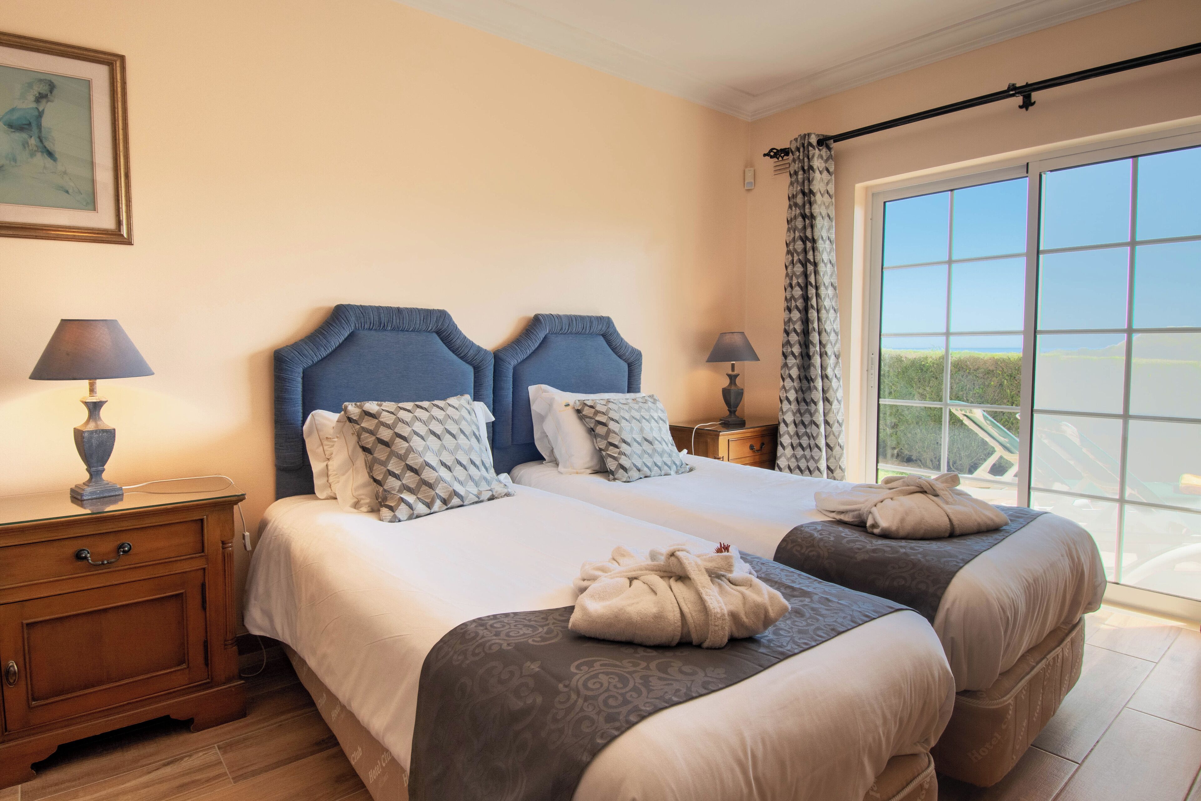 A hotel room with two beds, a wooden nightstand, a lamp, and a view of the outdoors.