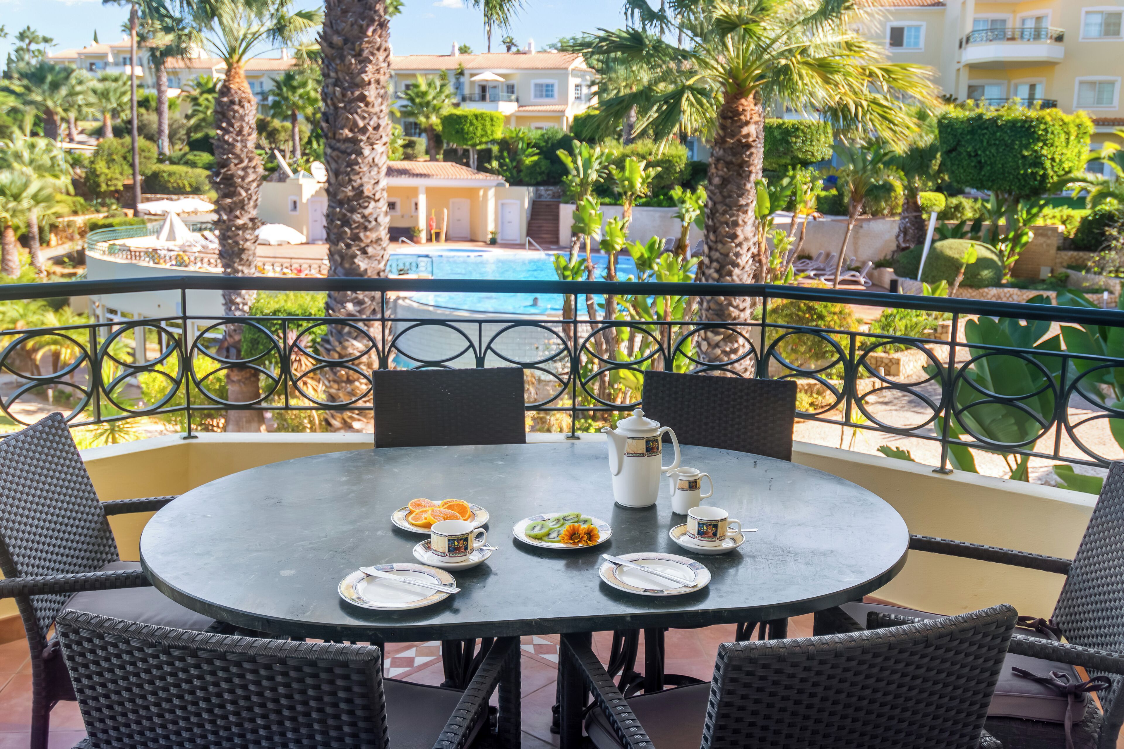 Une table ronde, sur laquelle sont disposées des assiettes, des tasses et une carafe, se trouve sur un balcon donnant sur une piscine et des palmiers.
