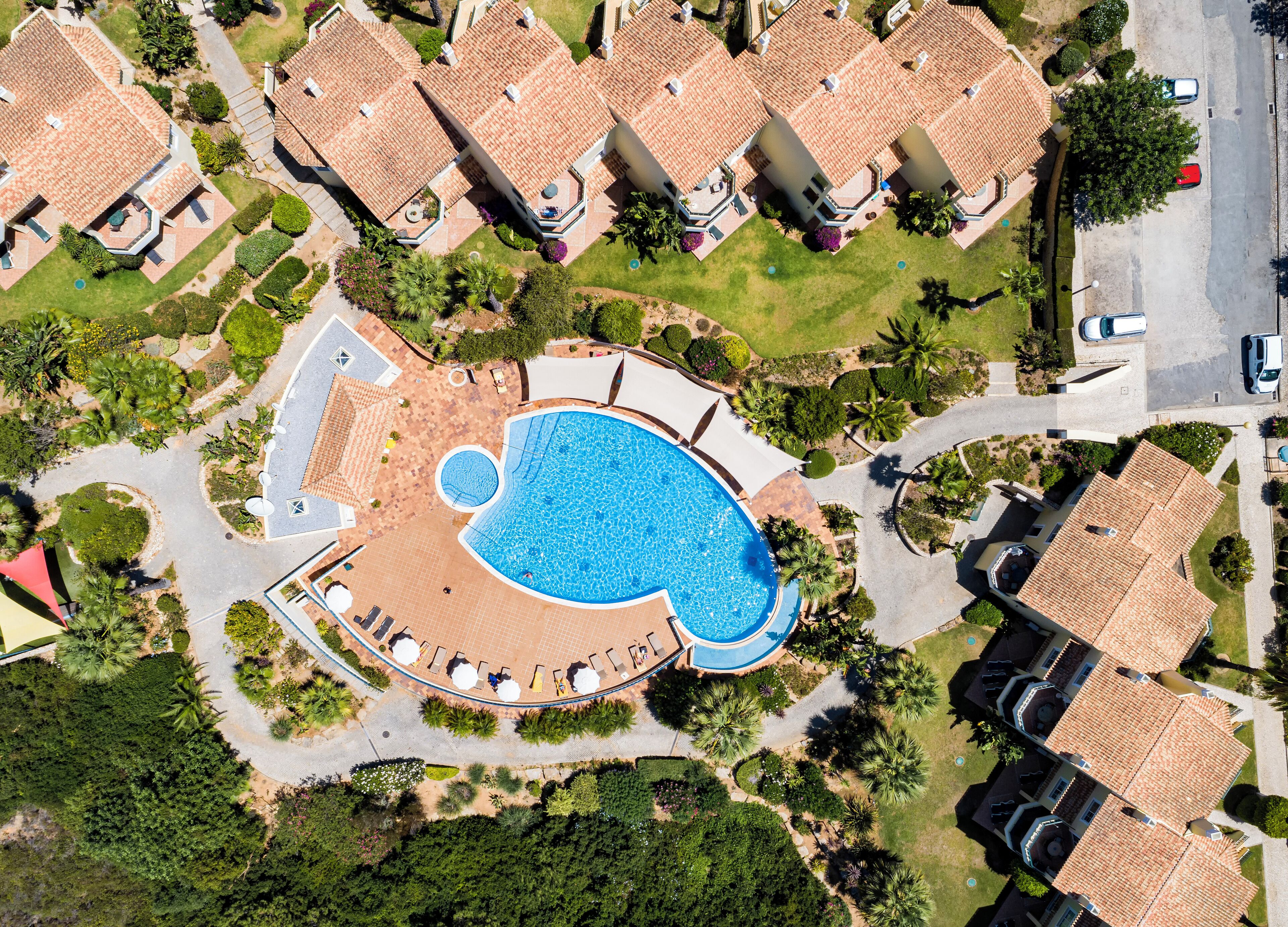 An aerial view of a residential area with a large swimming pool surrounded by a patio area with lounge chairs and umbrellas.