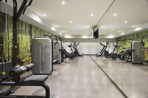 Uma academia com diversos aparelhos de exercícios, um espelho e um mural com tema de floresta na parede.