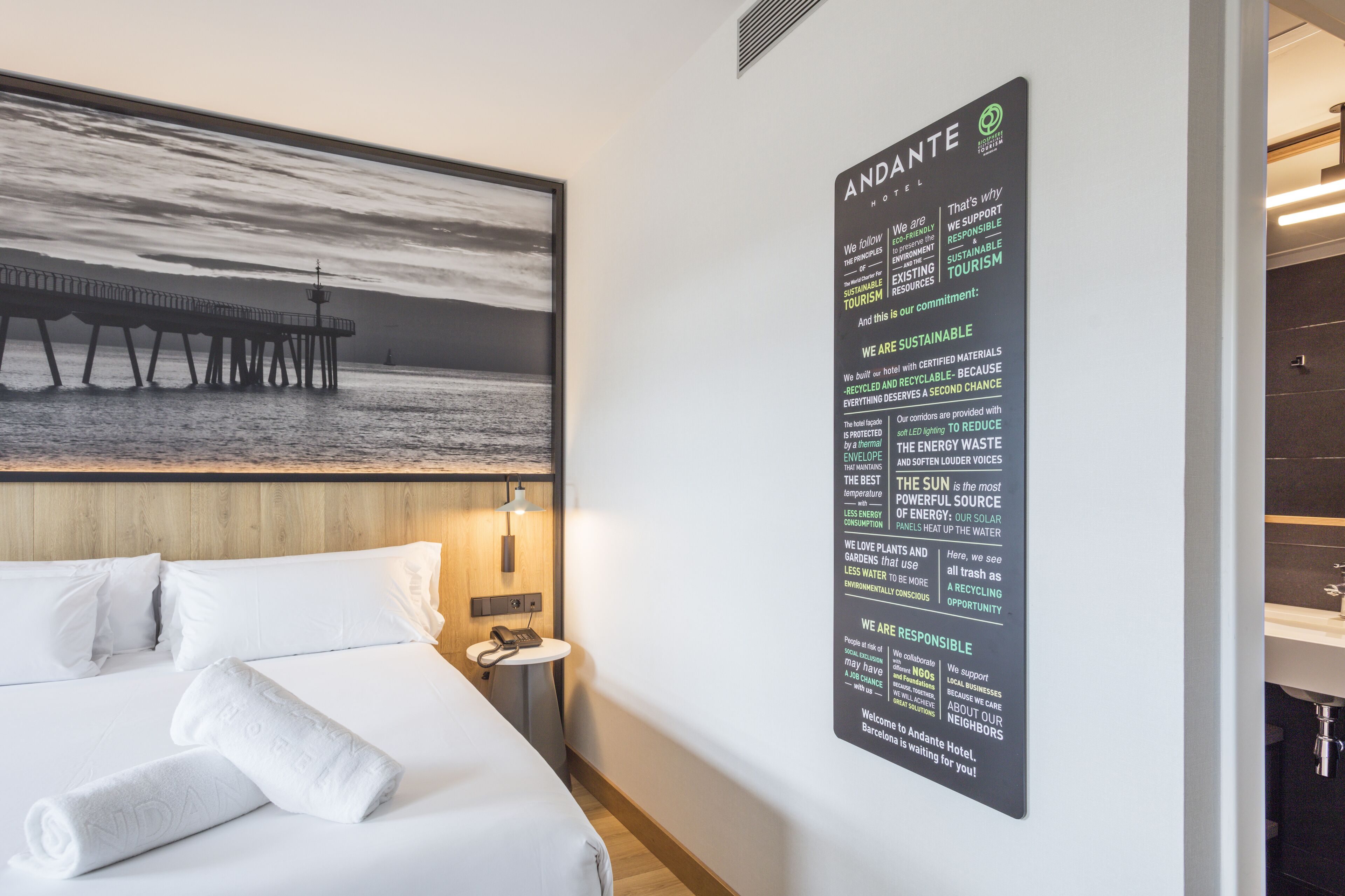 Habitació d'hotel amb un llit, una tauleta de nit, un cartell fixat a la paret que explica les iniciatives de sostenibilitat de l'hotel i una fotografia gran amb un moll.