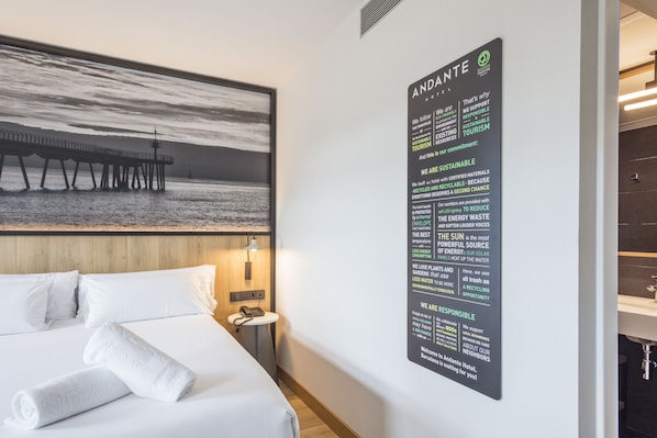 A hotel room with a bed, a nightstand, a wall-mounted sign detailing the hotel's sustainability efforts, and a large framed picture of a pier.