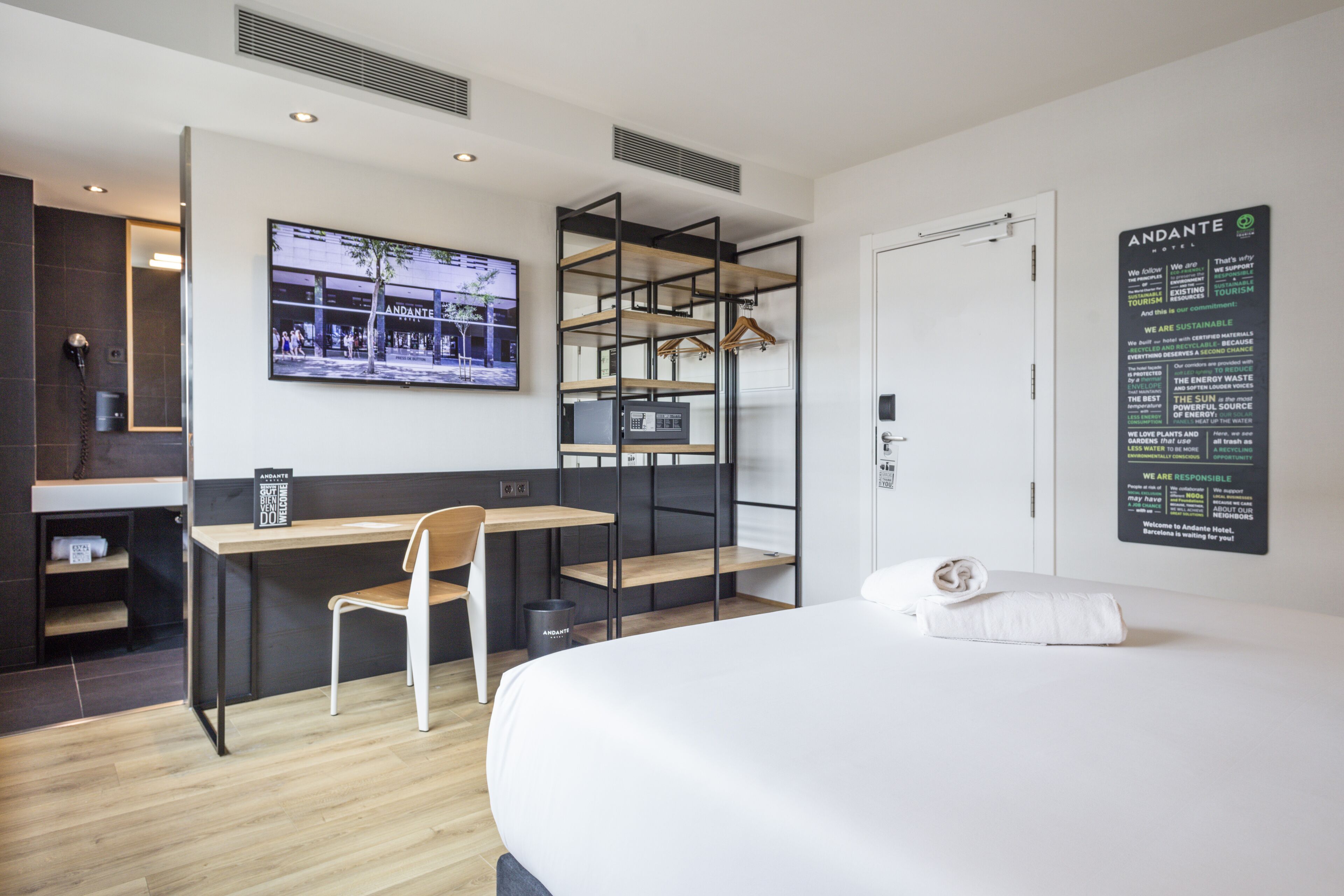 A hotel room with a bed, a desk, and a chair. There is a TV mounted on the wall, and a sign with the hotel's name and sustainability information.