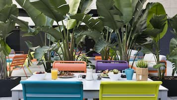 Un área de comedor al aire libre con sillas y mesas de colores, rodeada de grandes plantas tropicales.