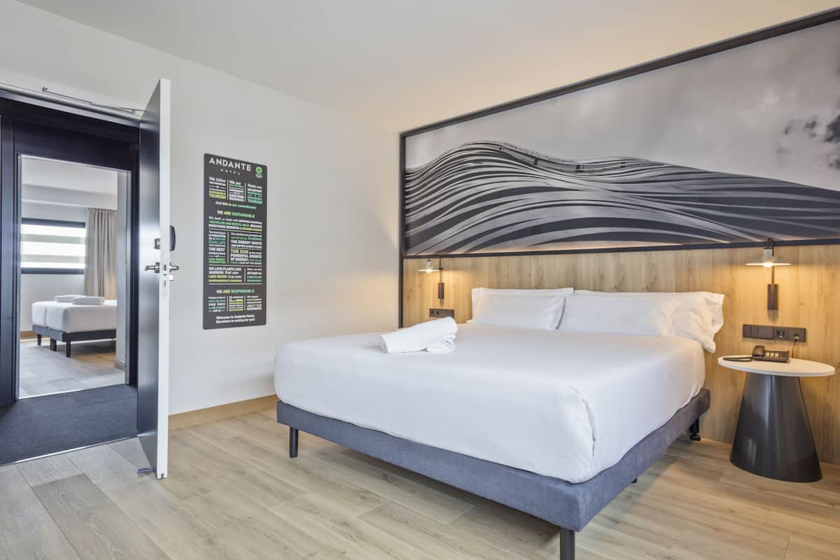 A modern hotel room with a large bed, a wooden headboard, a wall-mounted artwork, a bedside table with a lamp, and a poster on the wall.