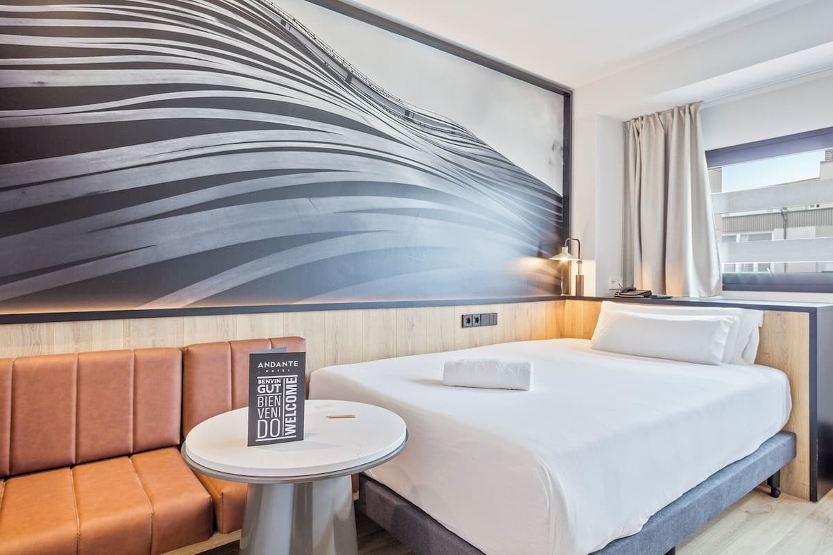 A hotel room with a bed, a round table, a brown leather sofa, a large abstract wall mural, and a window with curtains.