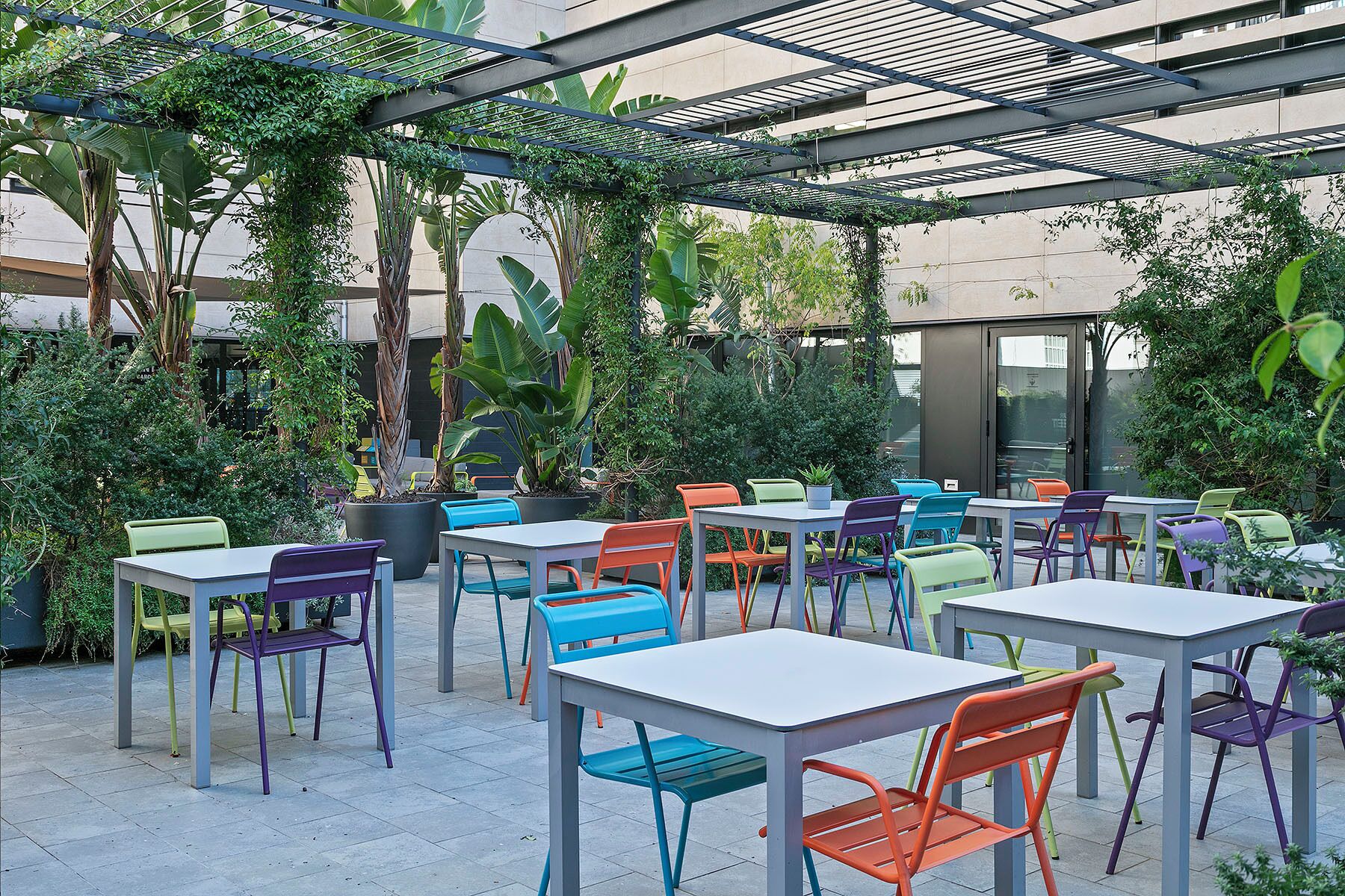 An outdoor patio area with tables and chairs, surrounded by greenery and a metal pergola structure.