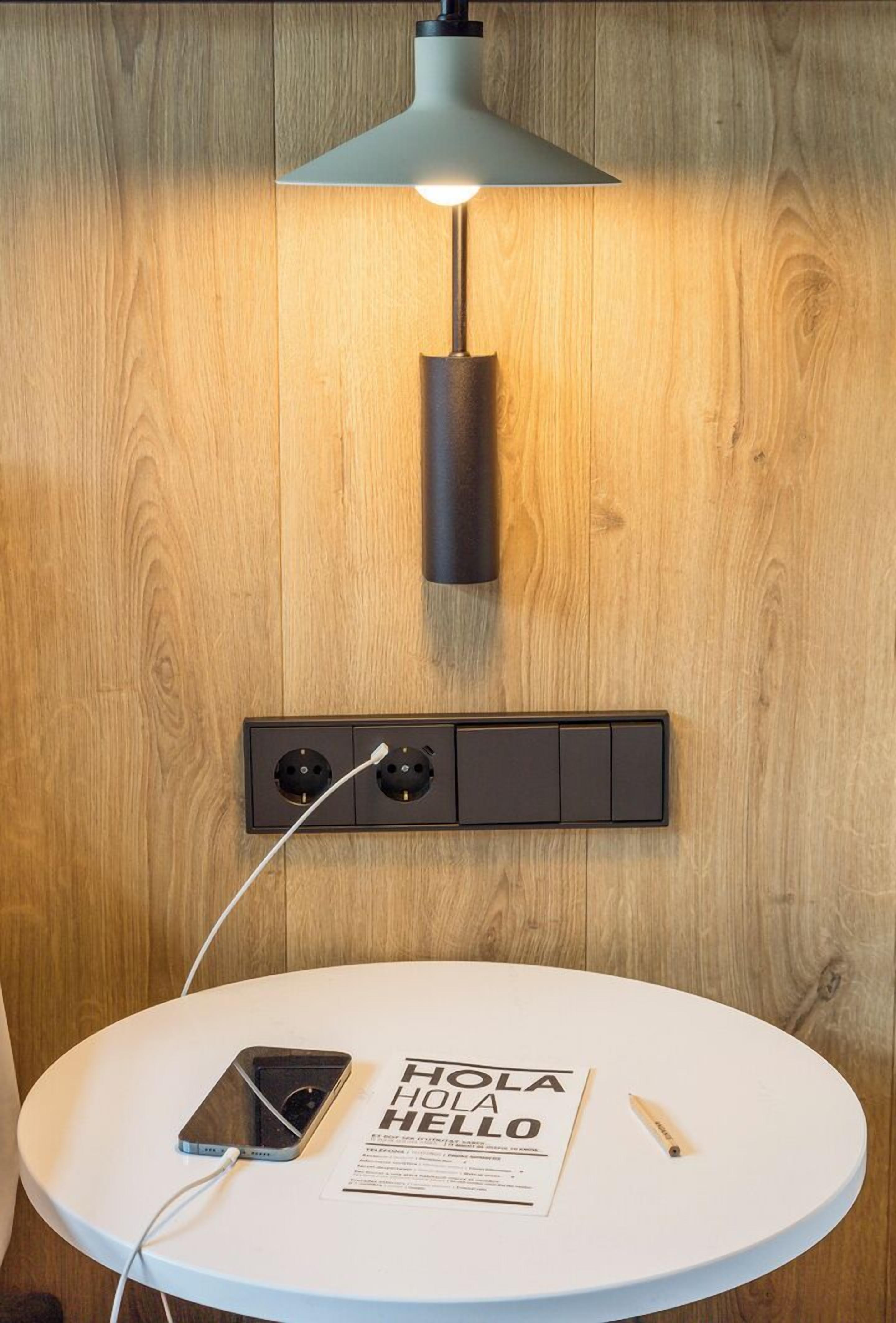 A modern room with a wooden wall, a round white table, a smartphone, a book titled "HOLA HELLLO", and a wall-mounted lamp.