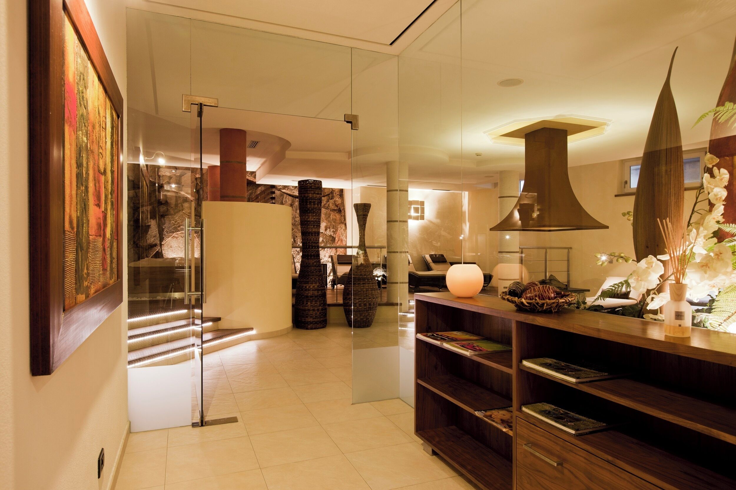 A modern hotel lobby with a glass partition, a staircase, a reception desk, and a decorative vase with flowers.