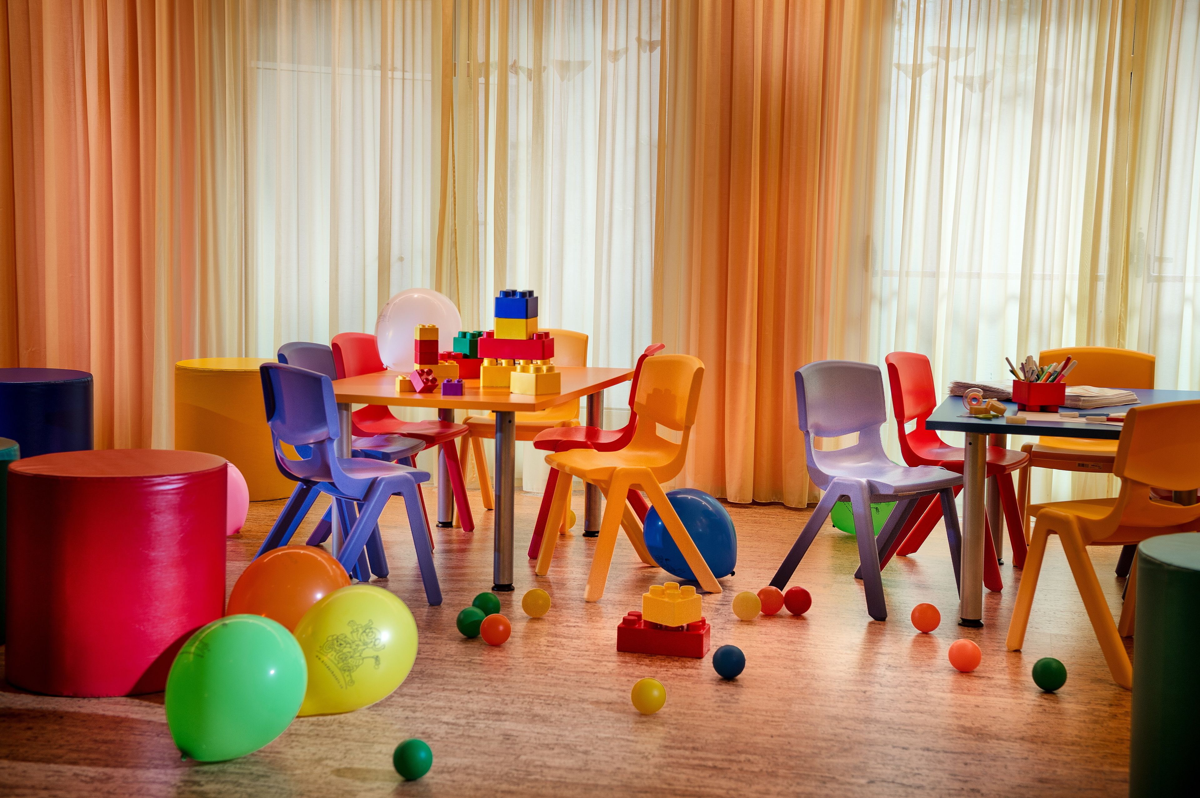 Een helder verlichte kamer met kleurrijke stoelen en een tafel, met diverse speelgoed en ballen verspreid op de vloer.