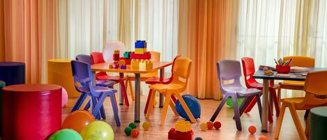 Een helder verlichte kamer met kleurrijke stoelen en een tafel, met diverse speelgoed en ballen verspreid op de vloer.