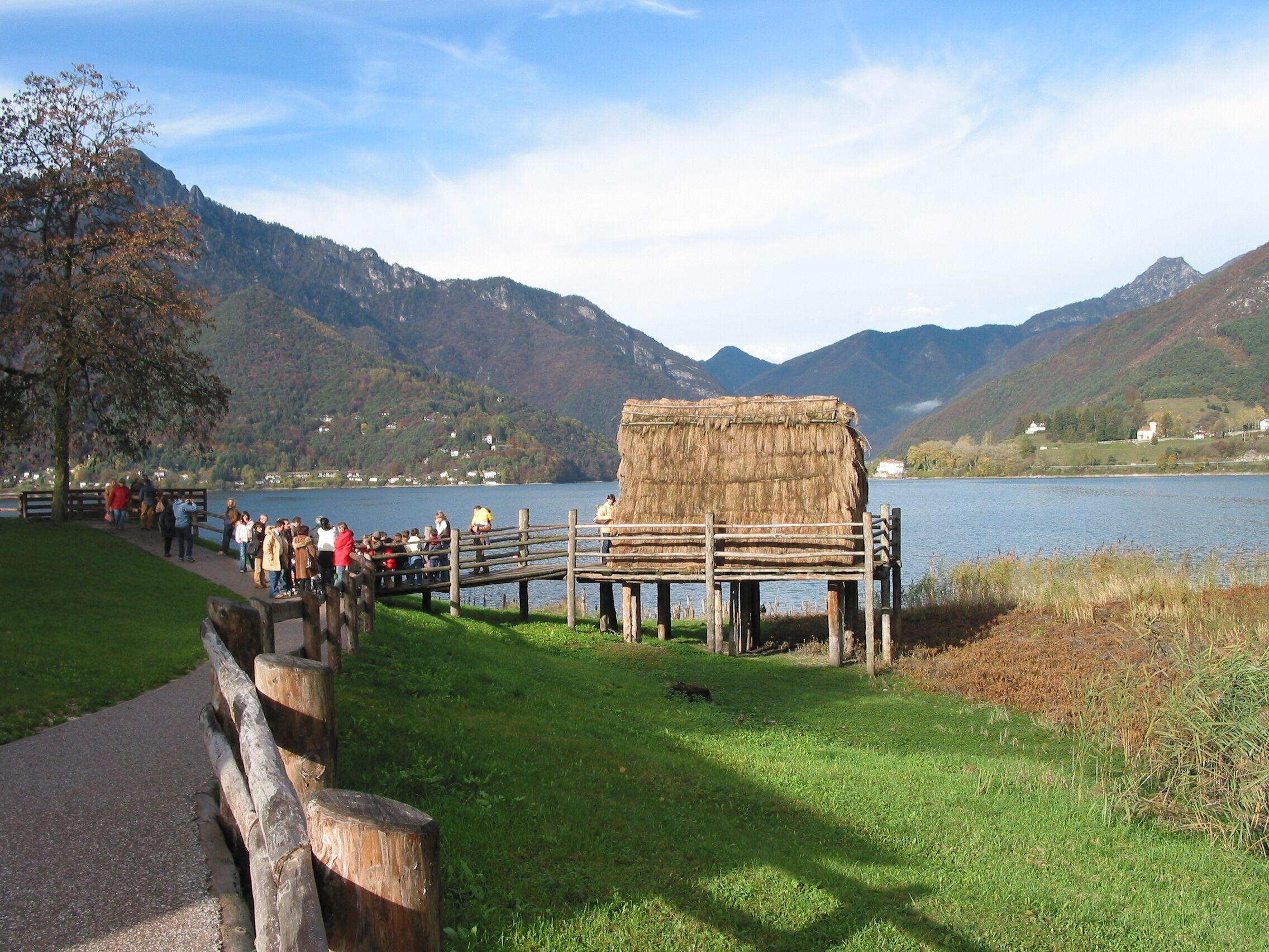 Une terrasse en bois surmontée d’une structure en chaume, un lac et des montagnes en arrière-plan.