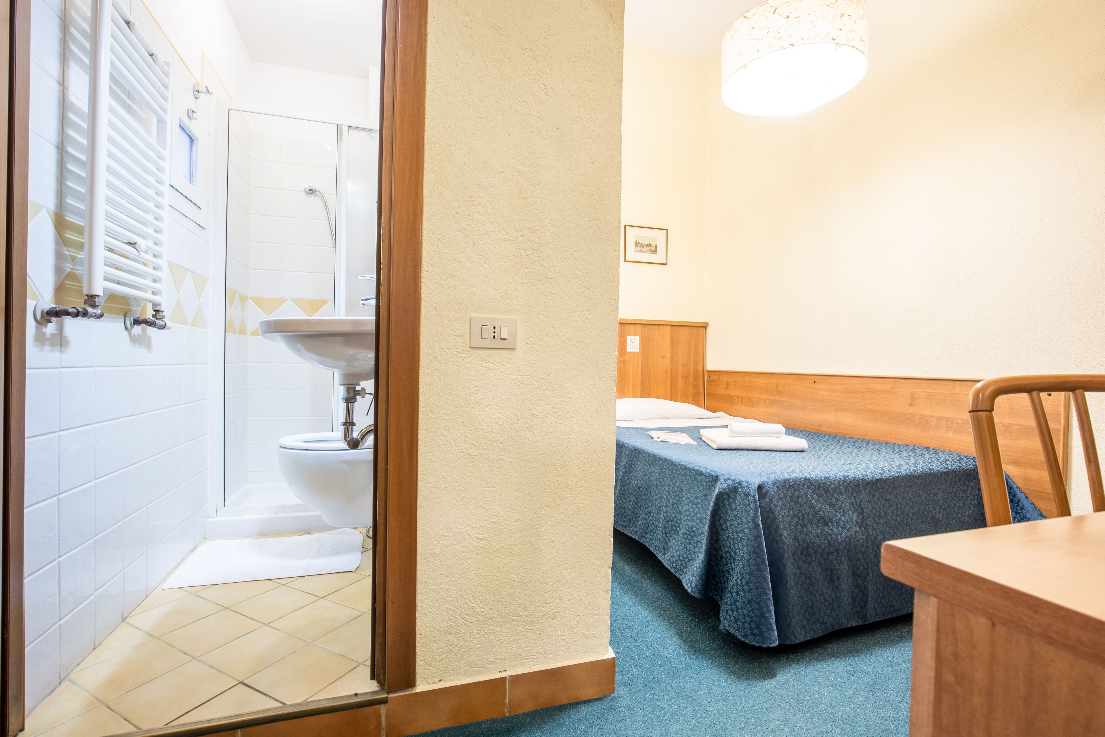 A bathroom with a shower, toilet, and sink. There is a bed with a blue blanket and a wooden headboard.