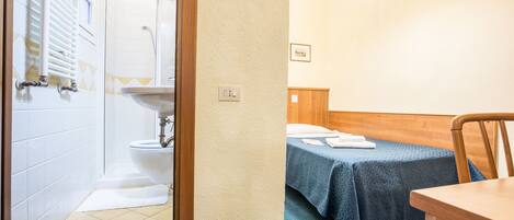 A bathroom with a shower, toilet, and sink. There is a bed with a blue blanket and a wooden headboard.