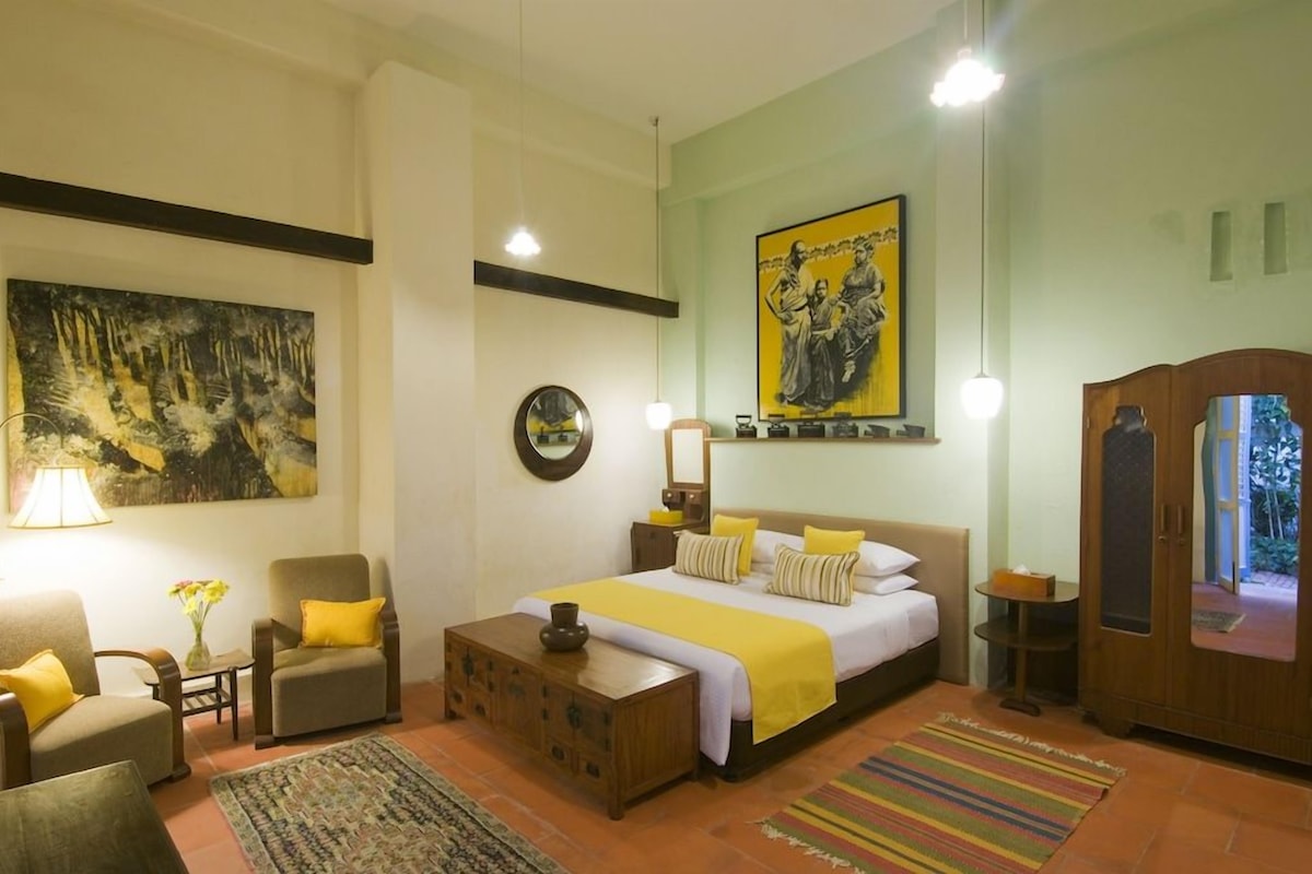 Habitación de hotel con una cama, dos sillones, un armario de madera, un espejo y obras de arte en las paredes.