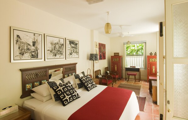 A neatly arranged bedroom with a large bed, a wooden headboard, and multiple pillows. There are framed artworks on the wall, a desk with a chair, and a red cabinet.