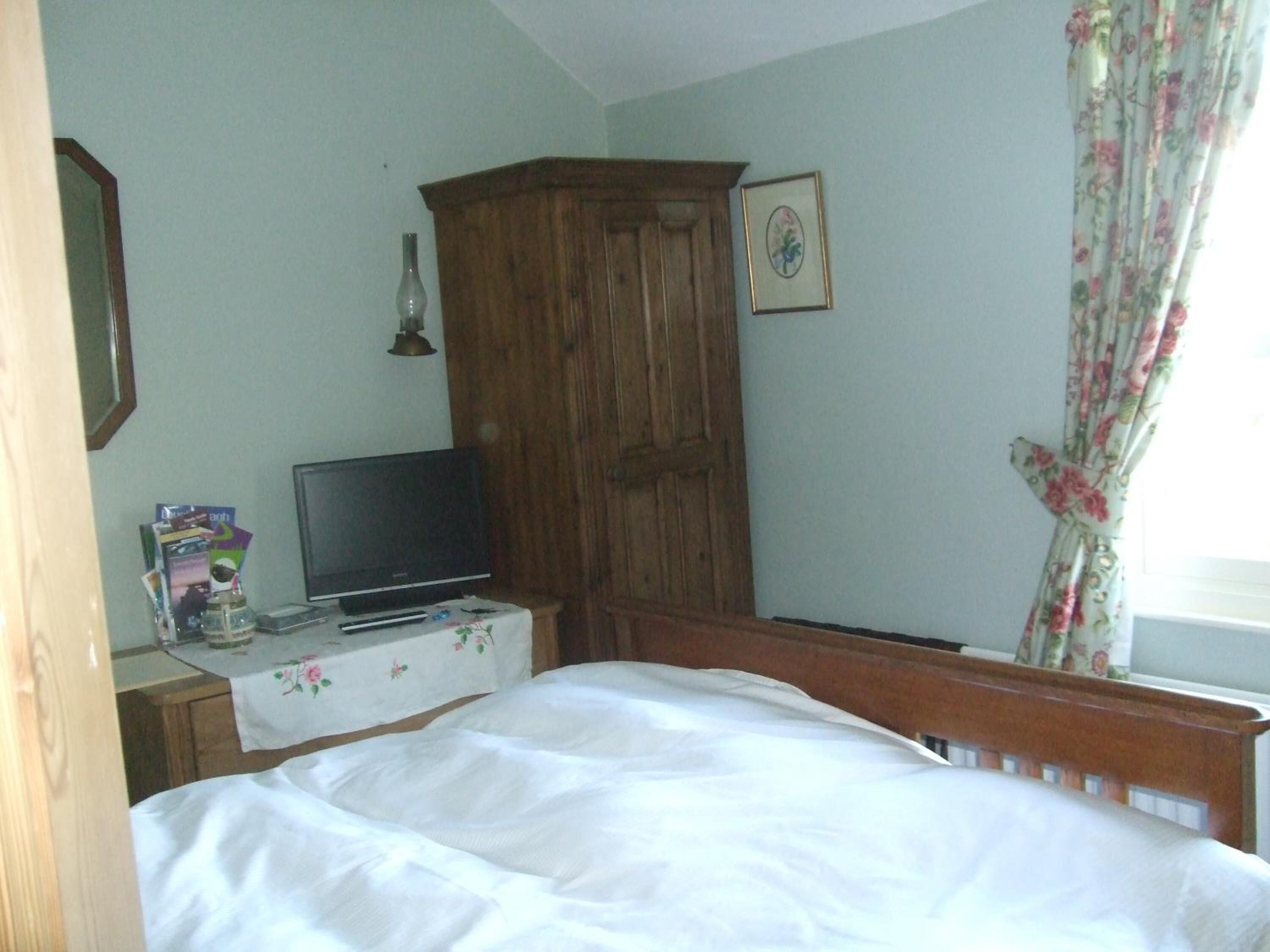 A bedroom with a bed, a wooden wardrobe, a television on a table, and a window with floral curtains.
