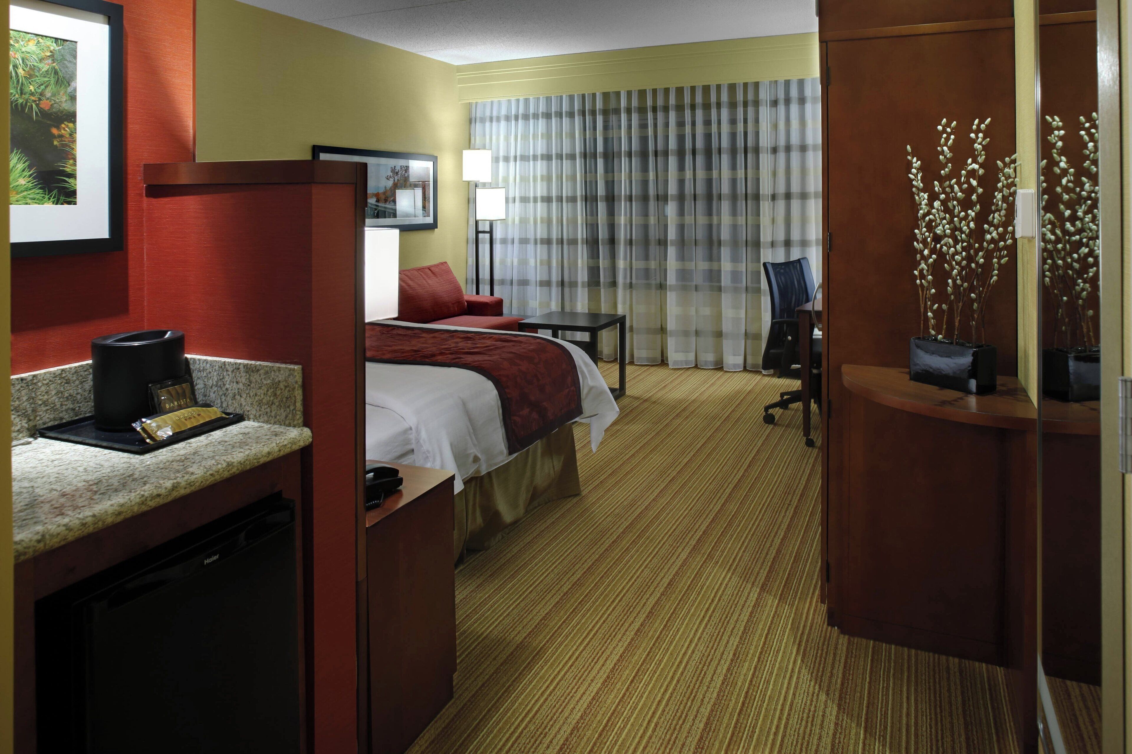 Una habitación de hotel con cama, escritorio con cafetera, silla, lámpara de escritorio y ventana con cortinas.
