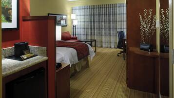 Una habitación de hotel con cama, escritorio con cafetera, silla, lámpara de escritorio y ventana con cortinas.