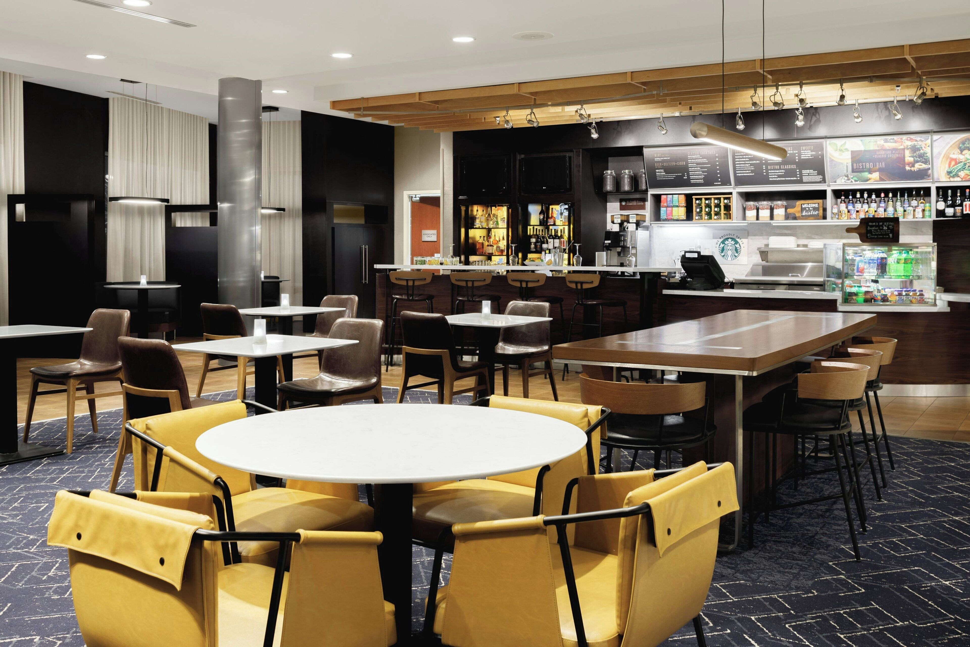 A modern restaurant with round tables and yellow chairs, a bar area with menu boards, and a coffee machine.
