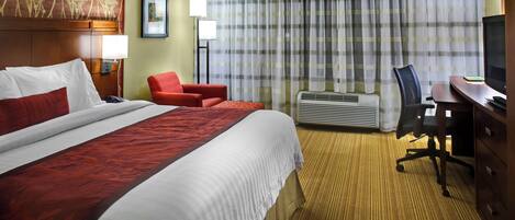 A hotel room with a large bed, a desk, a chair, a television, and an air conditioning unit.