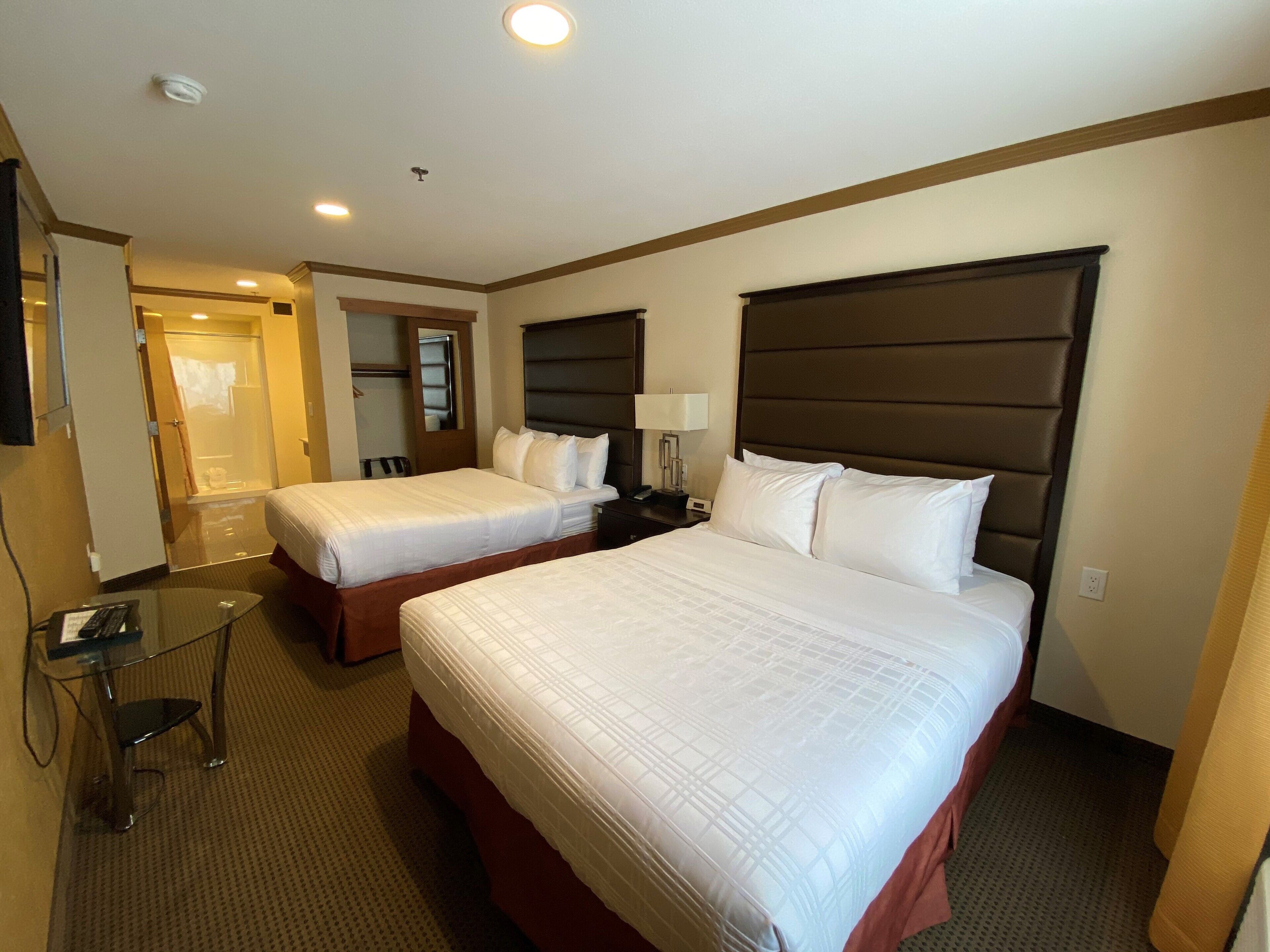 A hotel room with two beds, a desk with a chair, a mirror, and a shower area.