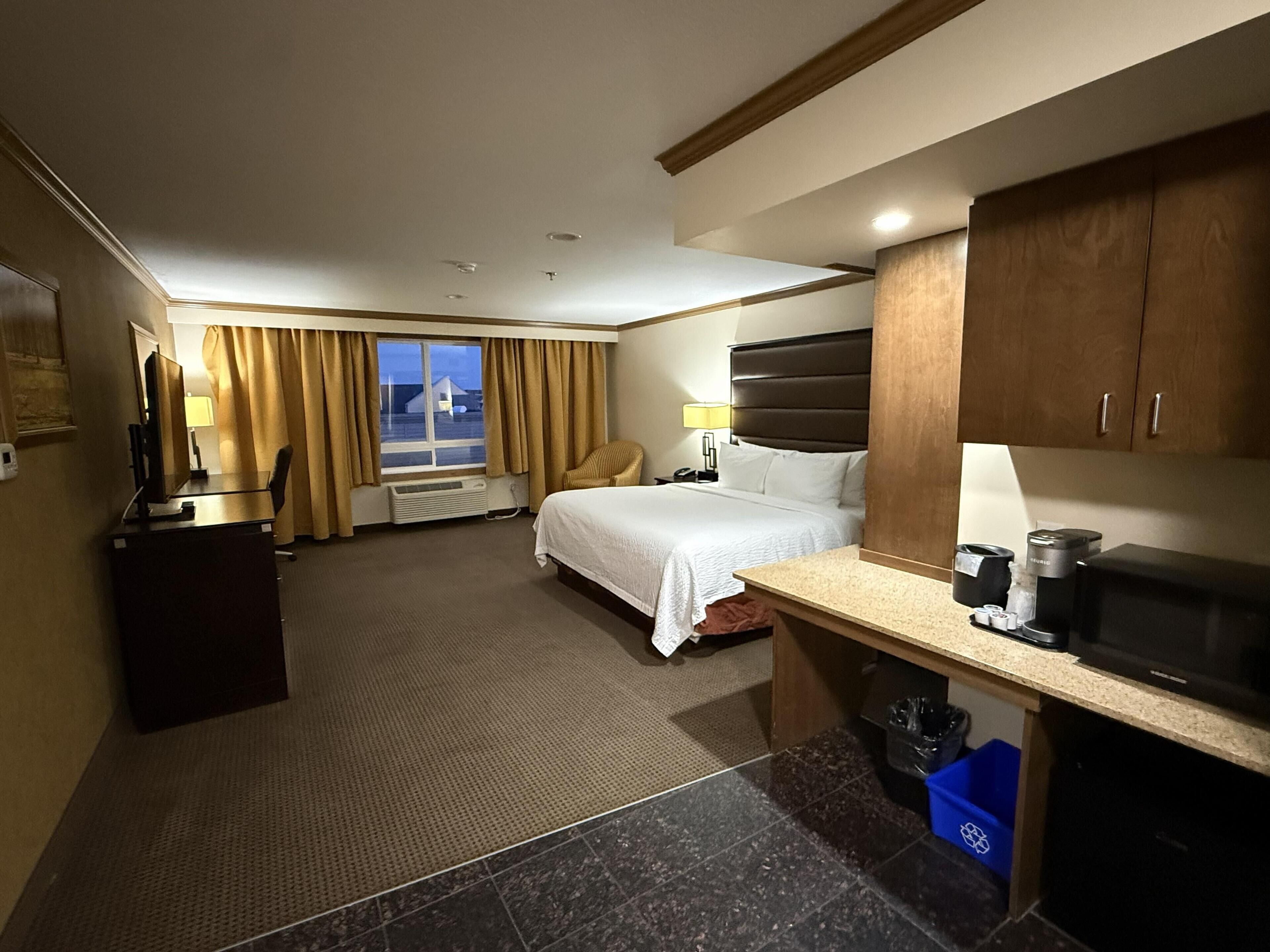 A hotel room with a bed, desk, kitchenette, and a window with curtains.