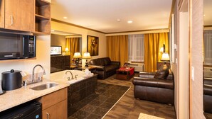 A hotel room with a kitchenette, a sofa, a coffee table, a TV, and a bathroom with a sink and mirror. - Best Western Plus Rose Country Hotel (Camrose)