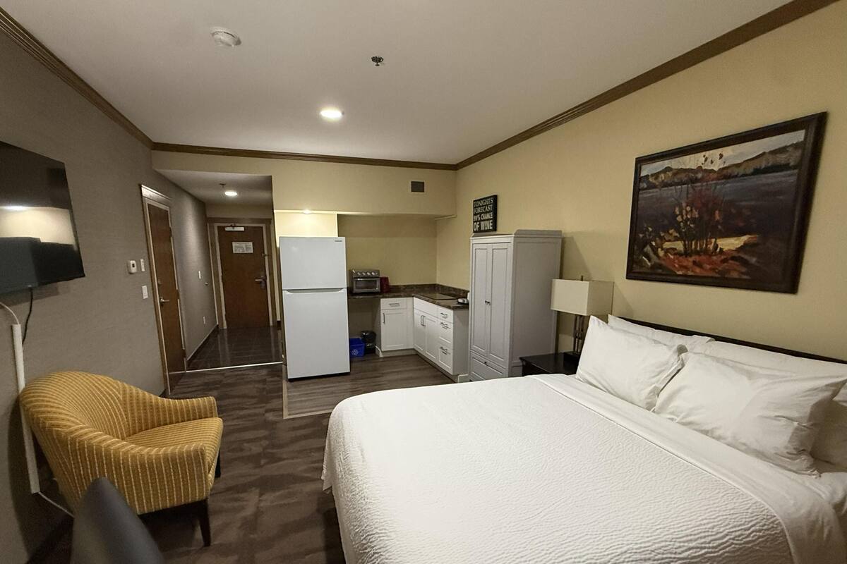 A hotel room with a bed, a chair, a kitchenette, and a painting on the wall.