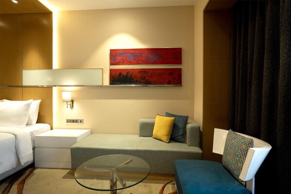 A hotel room with a bed, a sofa, a glass coffee table, and two wall-mounted artworks.