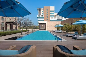 A swimming pool with lounge chairs and umbrellas, surrounded by a wooden deck and a building in the background. - Radisson Blu Hotel Amritsar (Amritsar)