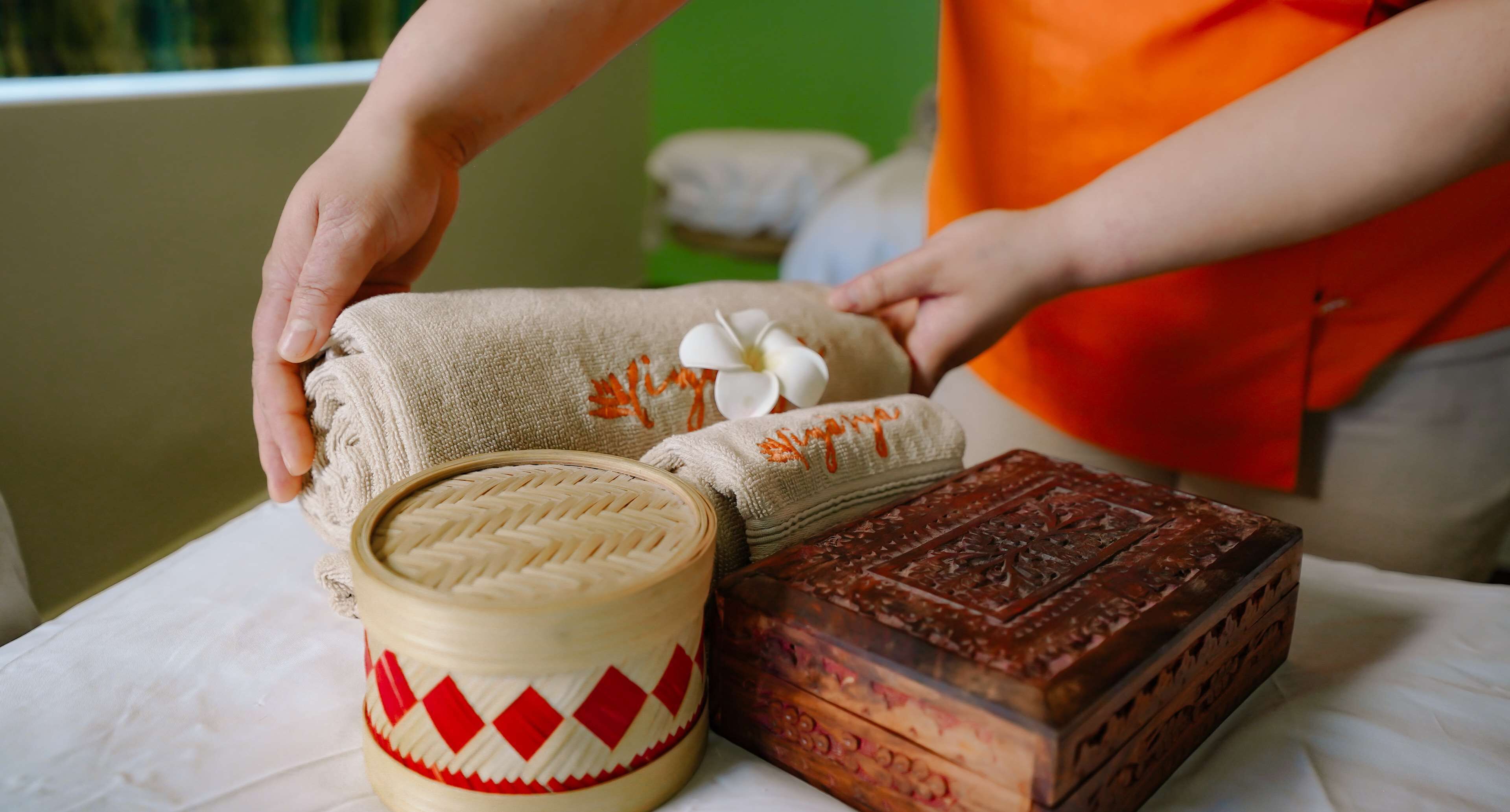 Uma mão colocando uma toalha bege com padrões vermelhos e brancos sobre uma mesa ao lado de uma caixa de madeira com entalhes detalhados.