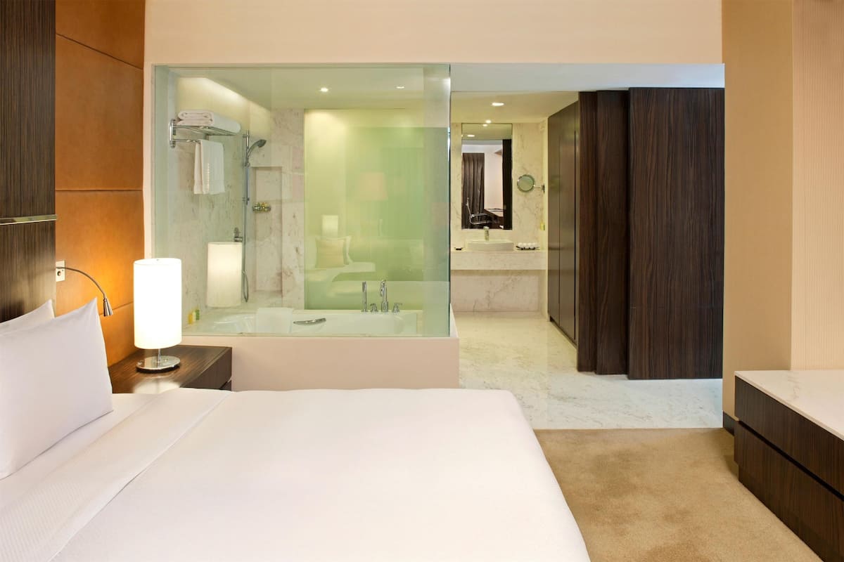 A modern hotel room with a large bed, a glass-enclosed shower area, a separate bathroom with a sink and mirror, and wooden paneling on the walls.