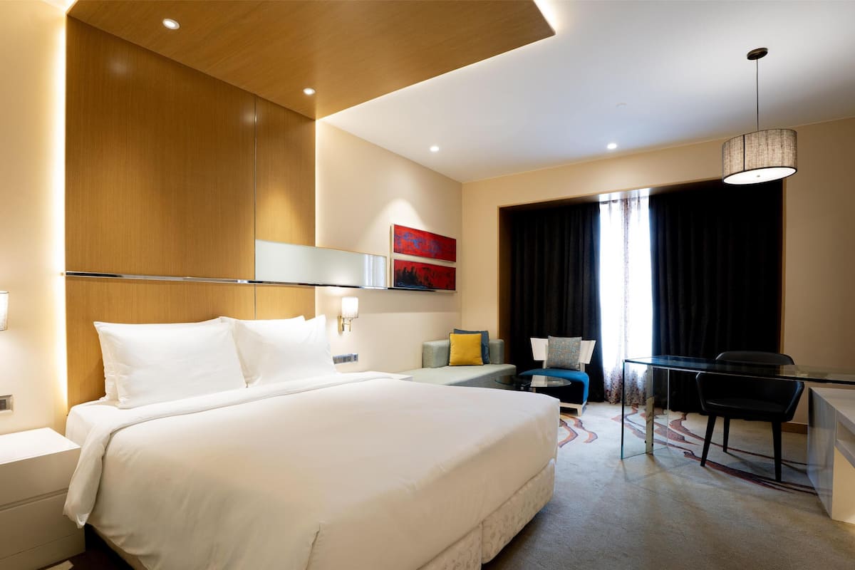 A modern hotel room with a large bed, a desk with a chair, a desk lamp, and a large window with curtains.