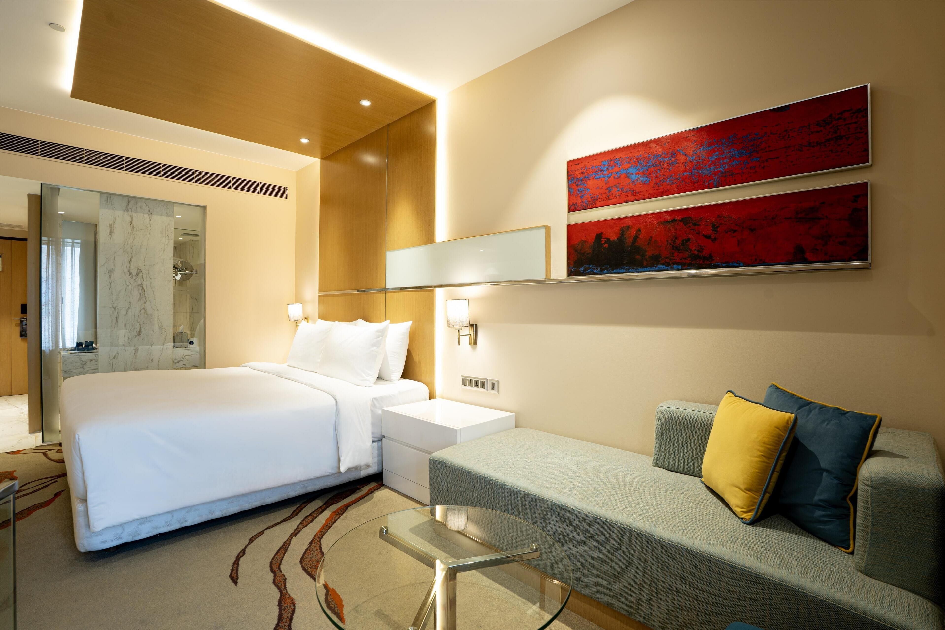 A modern hotel room with a large bed, a sofa, a glass coffee table, and two wall-mounted artworks.