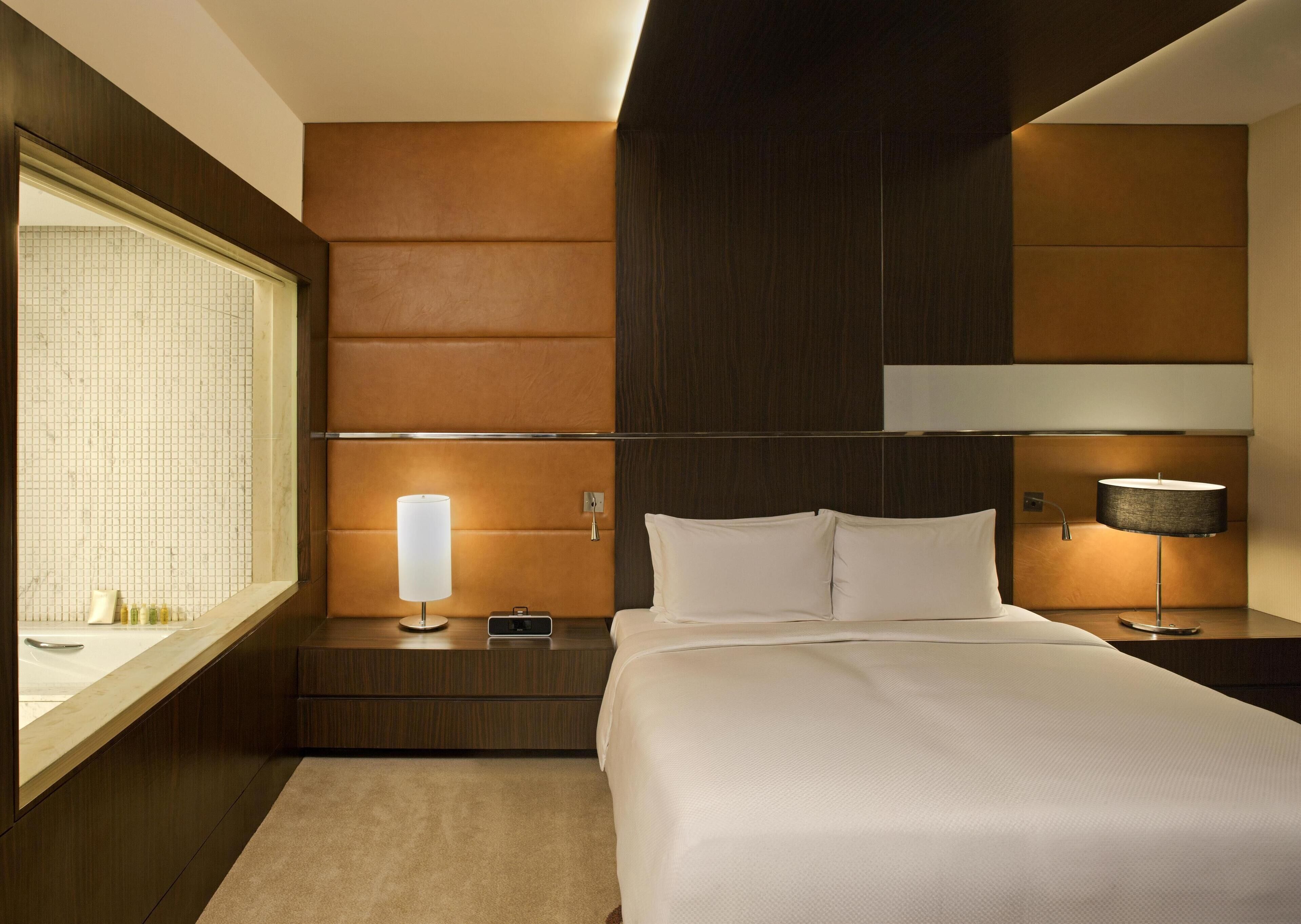 Una habitación de hotel moderna con una cama grande, una mesita de noche con lámpara, y una ventana con vistas a la ciudad.