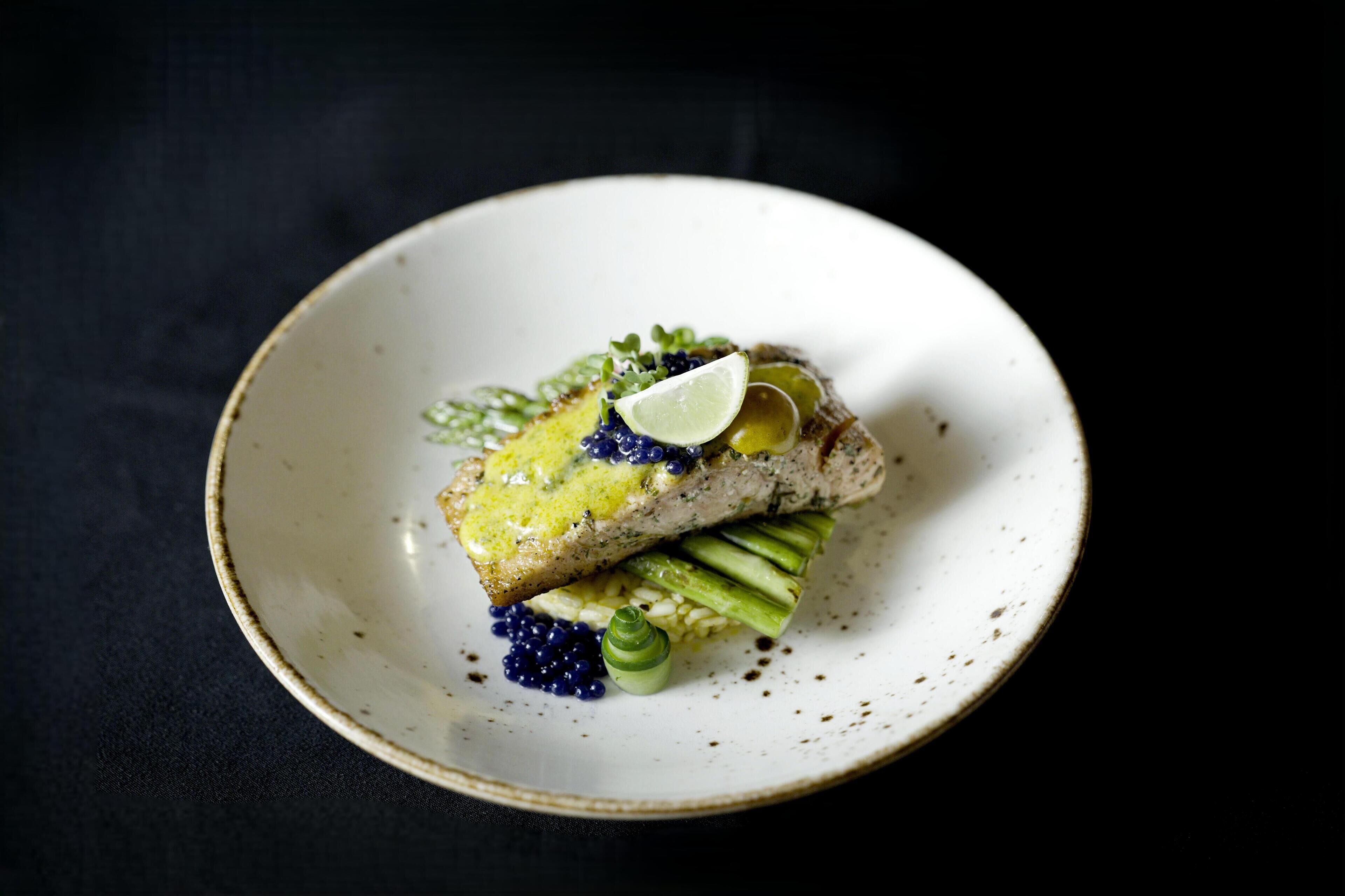 A plate with a piece of fish, green vegetables, and a slice of lime on a dark background.