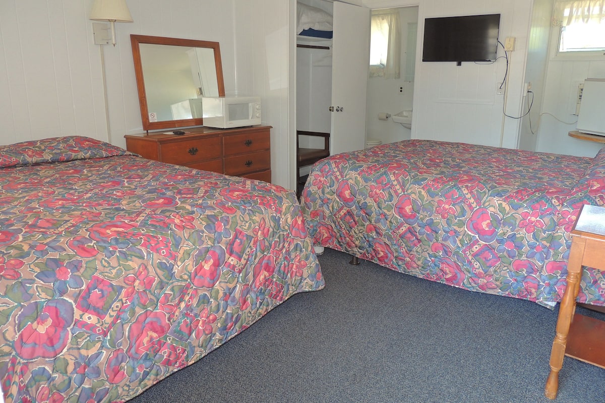 Habitación de hotel con dos camas, un aparador, un espejo, un televisor y una puerta que da a un baño.