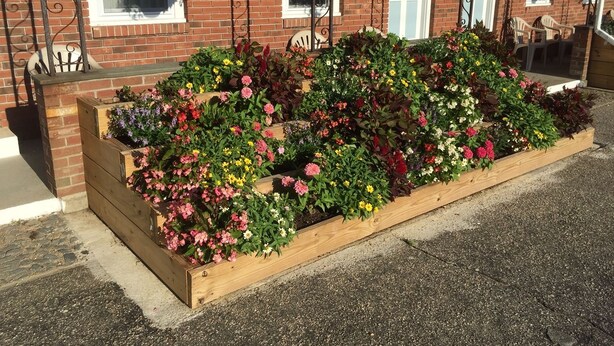Une jardinière en bois remplie d’une variété de fleurs aux couleurs vives, placée devant un bâtiment à la façade en briques et doté d’un porche avec des chaises.