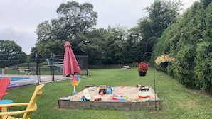 Une cour arrière clôturée avec un bac à sable, des jouets et un parasol rouge.