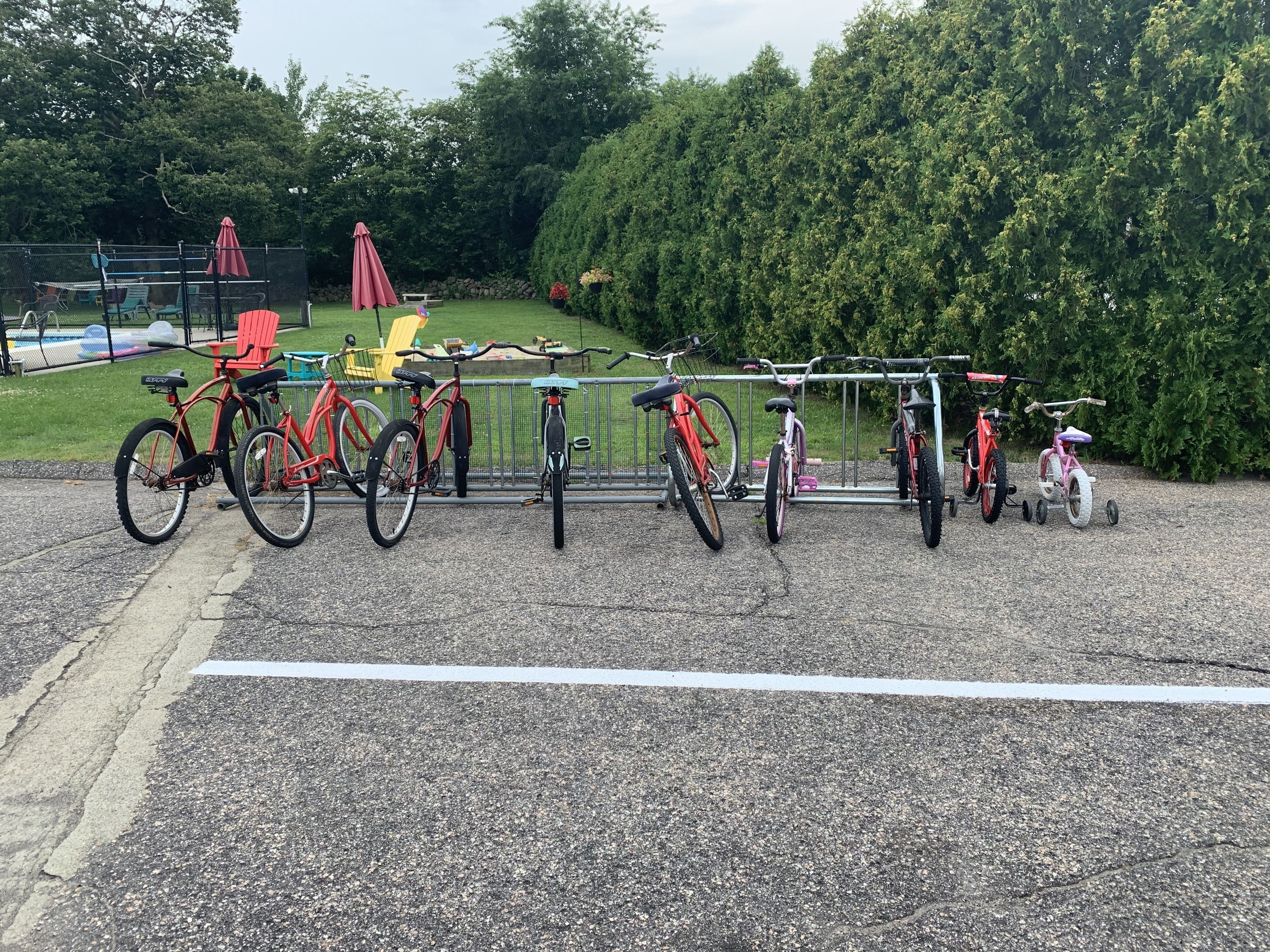 A row of bicycles parked in a gravel area with a metal fence and greenery in the background.