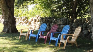 Une rangée de chaises Adirondack de différentes couleurs est disposée sur une pelouse, avec un mur de pierres et des arbres en arrière-plan.