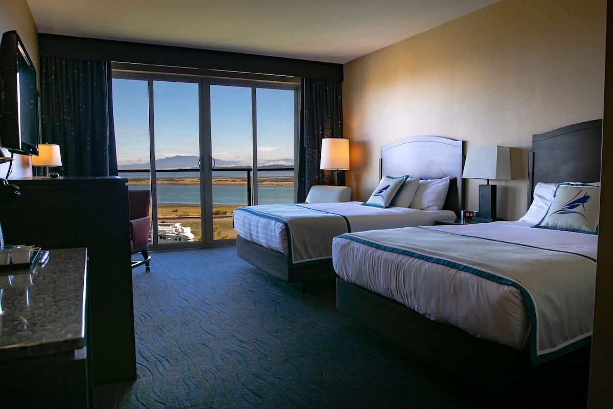 Habitación de hotel con dos camas, un escritorio, una silla y un ventanal con vista al mar.