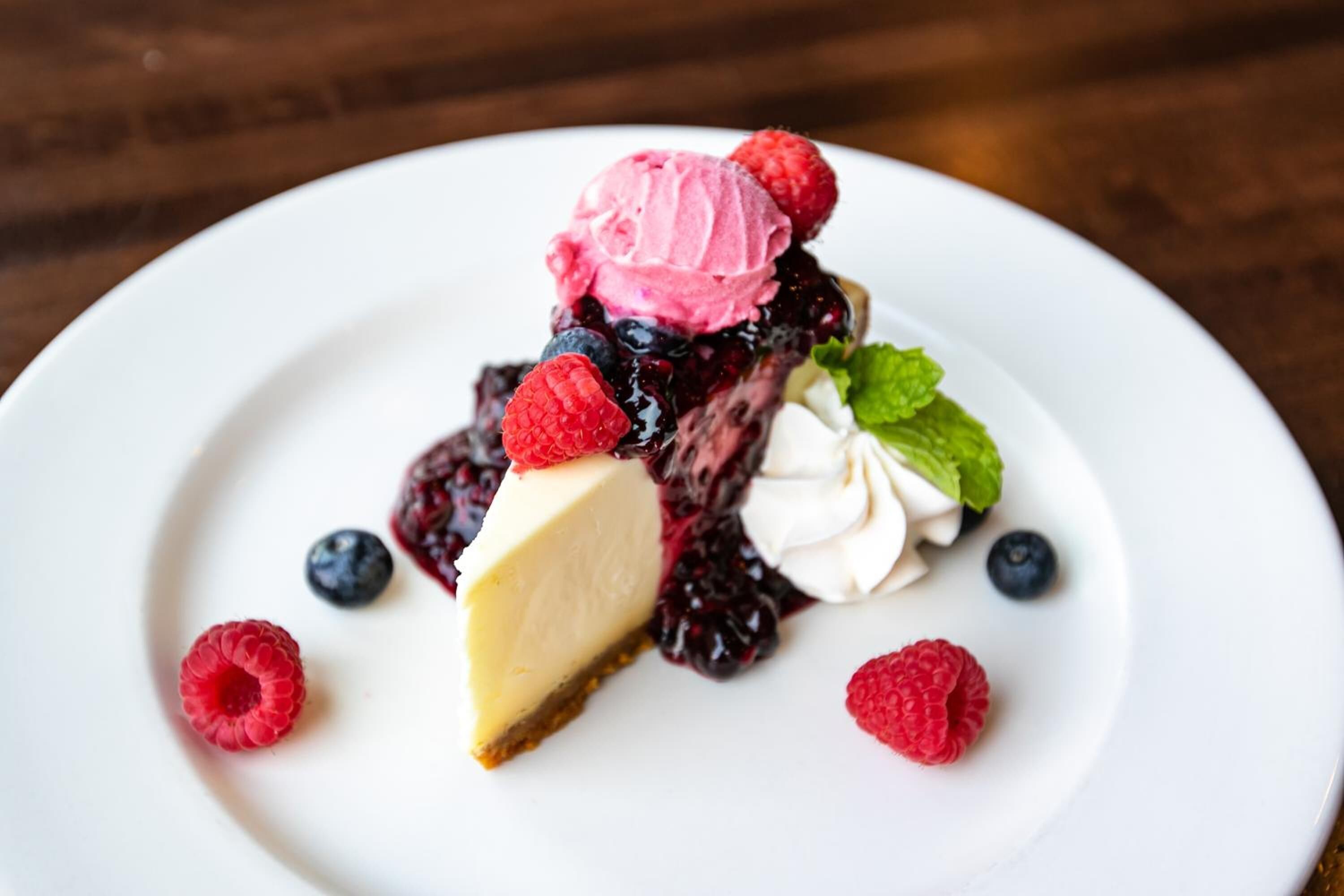 Un postre con una porción de pastel de queso, frutos rojos y una bola de helado sobre un plato blanco.