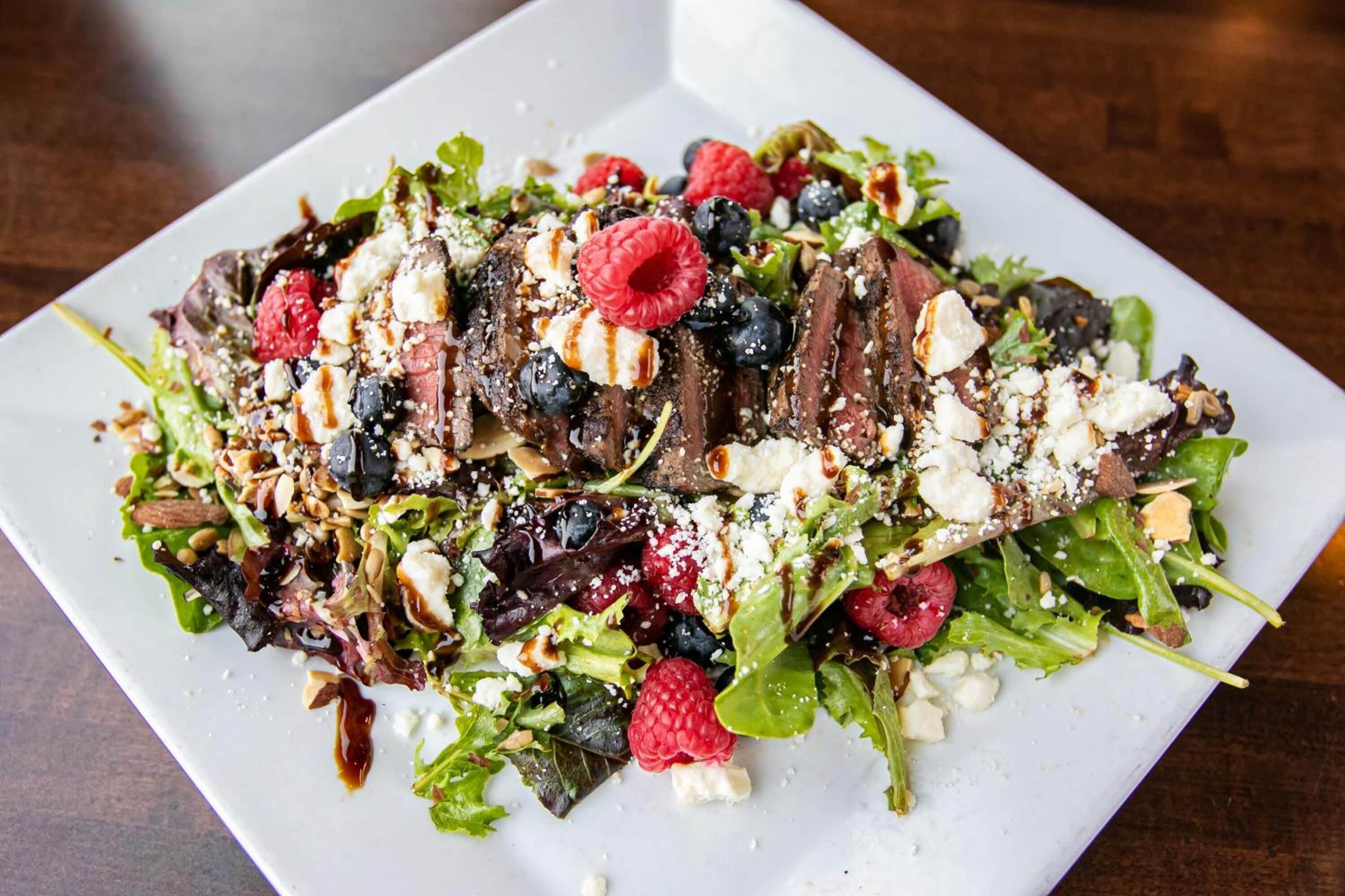 A white rectangular plate with a salad containing mixed greens, raspberries, blueberries, crumbled cheese, and chocolate shavings.